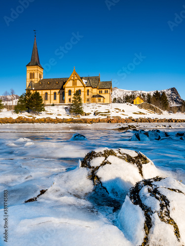 The wooden yellow Vågan Church on the beach on the Lofoten islands in Norway in winter with frozen river and mountains in morning light