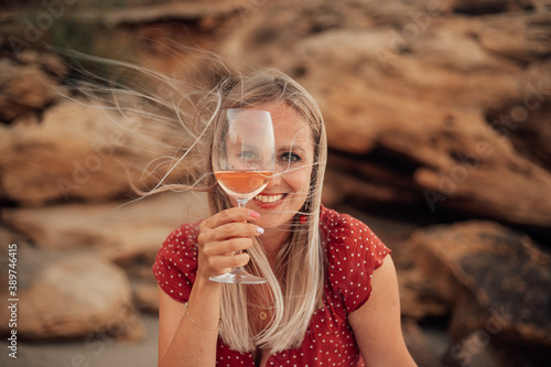 Beautiful happy blonde woman looks through a glass of wine. A woman sits in nature with a glass of wine.