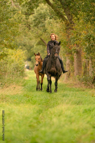 Young woman riding without saddle on her beautiful brown mare, yellow foal next to them, in the autumn forest