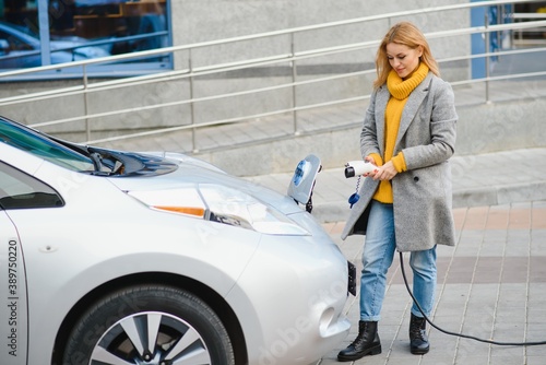 Stylish woman after buying products with a shopping bag is standing near the charging electric car