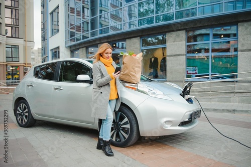 Charging electro car at the electric gas station. Woman standing by the car.