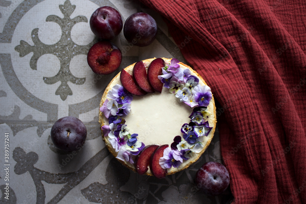 Cheesecake decorated with red plums and edible flowers. Fabric texture background. Top view photo of homemade dessert.
Cheesecake decorated with red plums and edible flowers. Fabric texture background. Top view photo of homemade dessert.