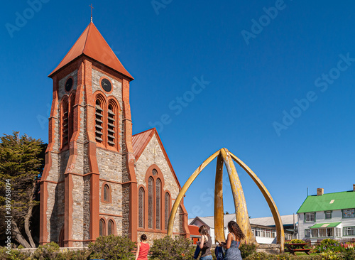 Stanley, Falkland Islands, UK - December 15, 2008: Closeup of Red and brown stone Christ Church Cathedral with tower under blue sky in its garden with green tree and whalebone arch.. Green roof adds c photo