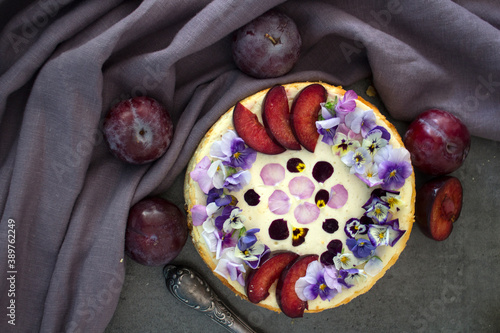 Cheesecake decorated with red plums and edible flowers. Fabric texture background. Top view photo of homemade dessert. 
