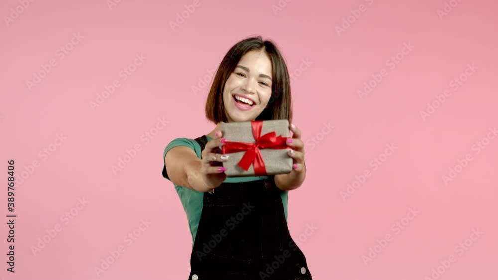 Excited woman holding gift box and gives it by hands to camera on pink wall background. Girl smiling, she is happy with present. Studio portrait Excited woman holding gift box and gives it by hands to camera on pink wall background. Girl smiling, she is happy with present. Studio portrait
