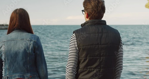 Young couplr go to sea along waterfront. Guy hugs girl standing by sea. Romantic date. View from the back. photo