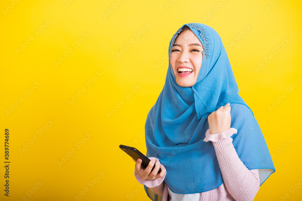Asian Muslim Arab, Portrait of happy beautiful young woman Islam religious wear veil hijab funny smile she raise hand glad celebrating winning with smart mobile phone isolated on yellow background Asian Muslim Arab, Portrait of happy beautiful young woman Islam religious wear veil hijab funny smile she raise hand glad celebrating winning with smart mobile phone isolated on yellow background