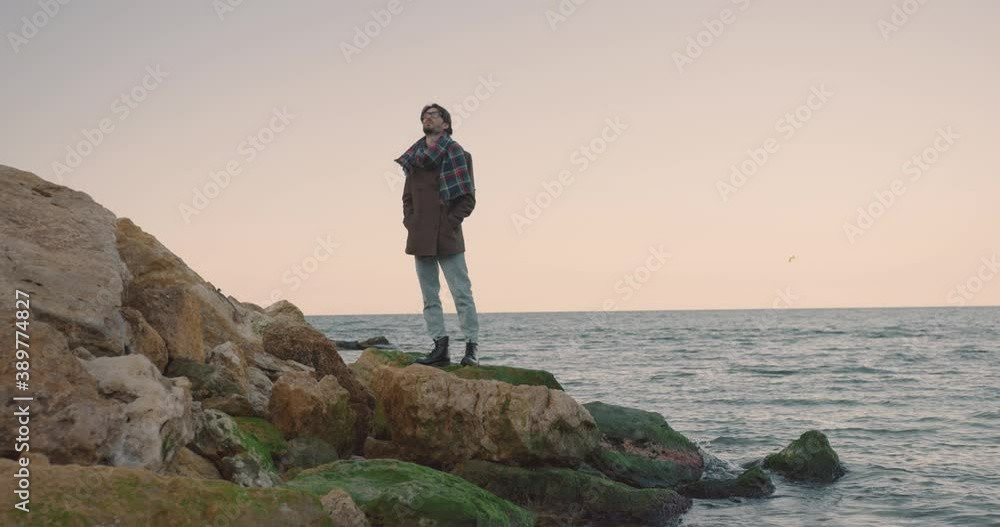 Сamera revolves around lonely young stylish man in autumn coat and glasses standing on rock by the sea. Сamera revolves around lonely young stylish man in autumn coat and glasses standing on rock by the sea.