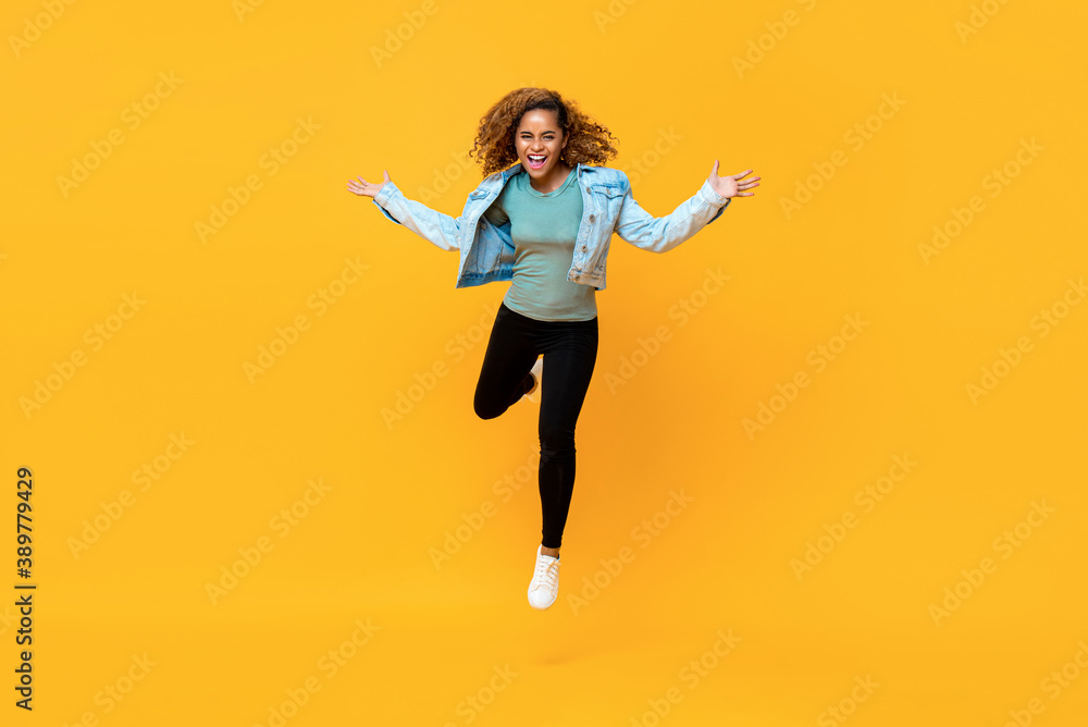 Full length portrait of energetic young African-American woman jumping in mid air while arms outstretched in  isolated studio yellow background