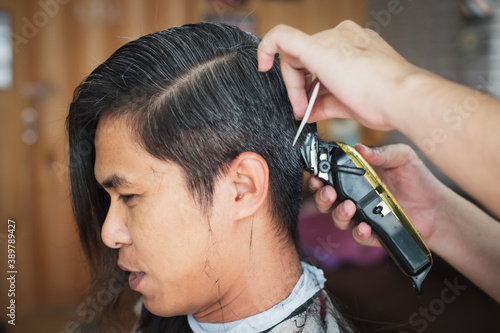 Asian young man being cut from long hair to short hair with electric clipper machine by professional barber in barbershop.