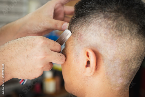 Fototapeta Naklejka Na Ścianę i Meble -  Asian young man being haircut with razor by professional barber in barbershop.