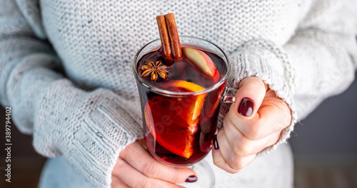 A woman in a white warm sweater is holding a transparent glass cup of mulled wine in her hand. Concept of a cozy atmosphere and a traditional winter drink