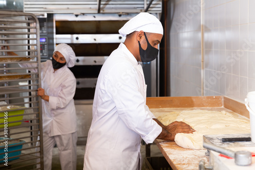 Worker of bakery in protective mask preparing fresh baked goods for sale on counter