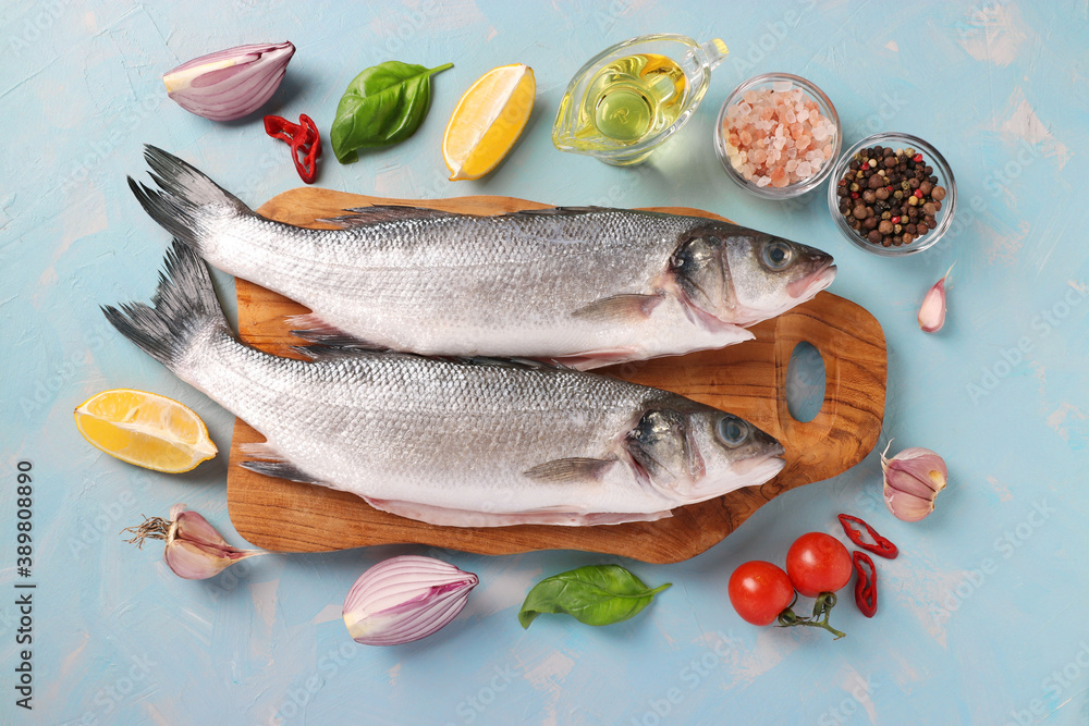 Raw fish seabass with ingredients and seasonings like basil, lemon, salt, pepper, cherry tomatoes and garlic on wooden board on light blue background. View from above