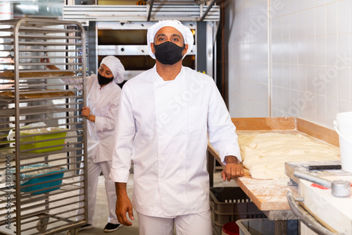 Worker of bakery in protective mask preparing fresh baked goods for sale on counter