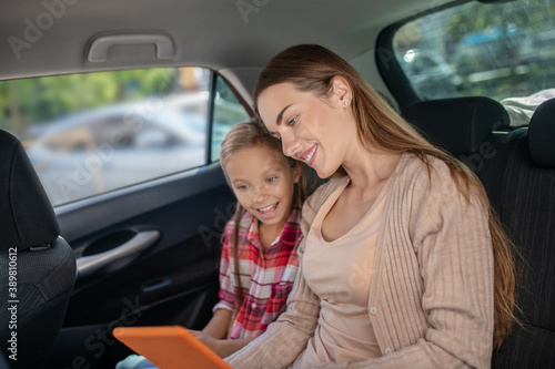 Pleased daughter and her mom checking something on tablet on backseat of car