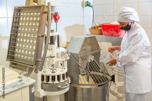 Young female bakery worker controlling dough kneading on professional equipment