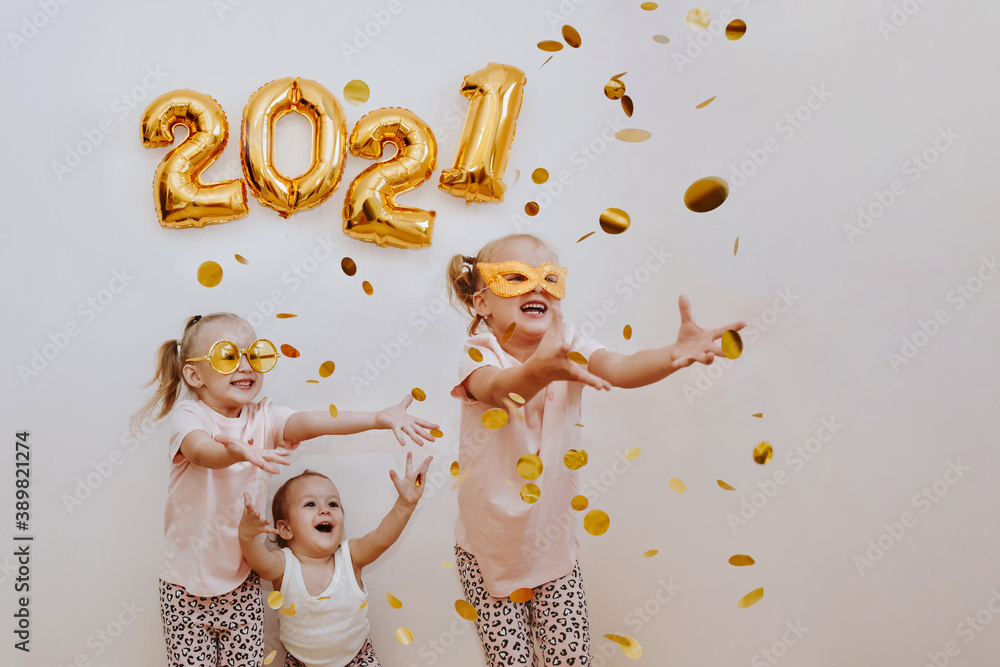 Three little girls are enjoying the flying golden confetti. Celebrating at home 2021. Concept for Christmas and New Year. - obrazy, fototapety, plakaty 