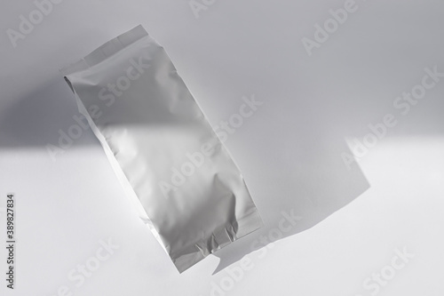 Blank white foil pouch bag on grey background in minimal style with natural harsh shadows. Monochrome food photo. Packaging valve and seal template mockup. Metallic coffee tea retail package design.