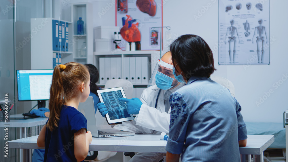 Radiologist explaining x-ray using tablet in medical office and nurse working on computer. Pediatrician specialist with protection mask providing health care service radiographic treatment examination