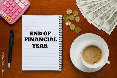 Flatlay picture of note book with end of financial year word, pen, milk coffee, fake cash, coins and calculator.