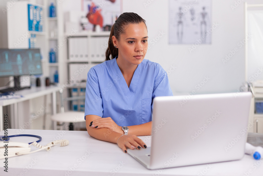 Medical nurse working on laptop in hospital office wearing blue uniform. Health care practitioner sitting at desk using computer in modern clinic looking at monitor, medicine. Medical nurse working on laptop in hospital office wearing blue uniform. Health care practitioner sitting at desk using computer in modern clinic looking at monitor, medicine.