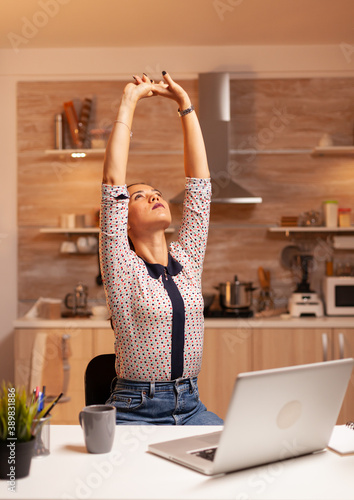 Exhausted freelancer woman streching while working on a deadline late at night. Employee using modern technology at midnight doing overtime for job, business, busy, career, network, lifestyle photo