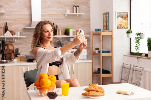 Sensual woman taking photos using smartphone in home kitchen while wearing sexy underwear. Seductive woman with tattoos using smartphone wearing temping underwear in the morning.