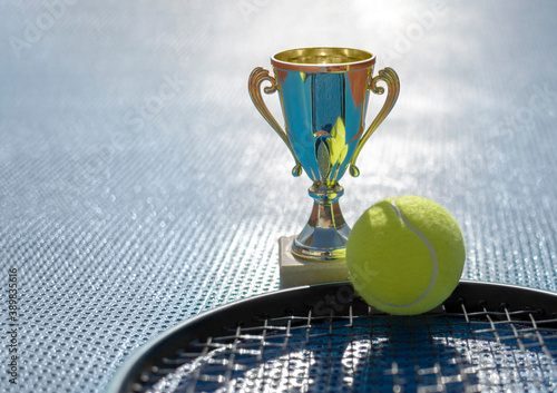 Gold champion trophy, tennis ball and racket on blue background in backlight. First place in tennis tournament. Award for the winner. Sports background, copy space for text © Elena