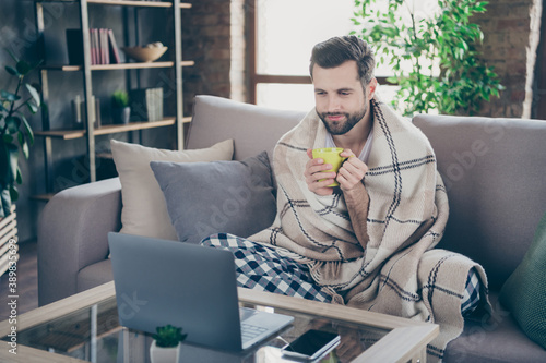Photo of handsome homey guy sit comfy sofa saturday weekend drink fresh coffee watch film notebook stay home quarantine pajama covered checkered blanket living room indoors