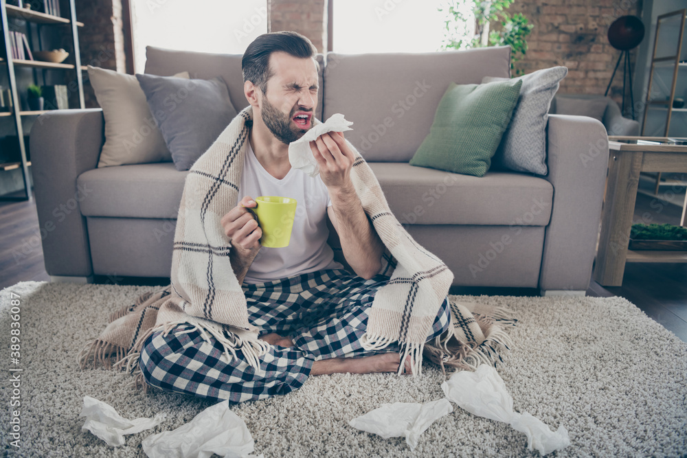 Obraz premium Full length photo of sick ill guy sit carpet hold hot tea beverage mug sneeze napkin cover plaid checkered blanket wear nightwear sleepwear in house indoors