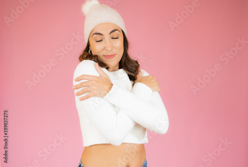 Portrait of a pretty young woman in winter clothes hugging herself standing isolated over pink background