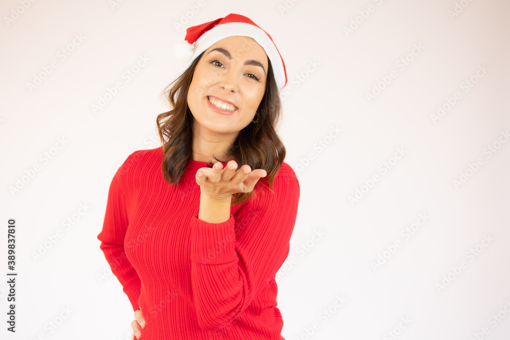Beautiful young woman wearing red sweater and santa hat sending air kiss over white background. Beautiful young woman wearing red sweater and santa hat sending air kiss over white background.