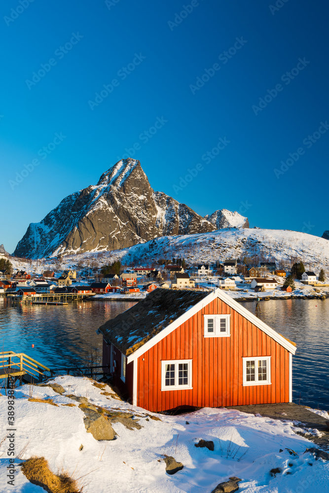 Renovated traditional red rorbu fishing hut on rocky coast near Reine on the Lofoten islands in Norway in winter with ocean and snow capped mountains in sunrise light