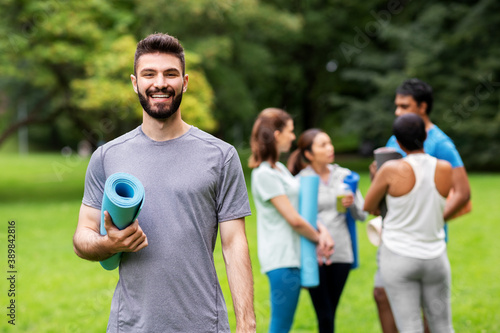 fitness, sport and healthy lifestyle concept - happy smiling young man with mat over group of people meeting for yoga class at summer park