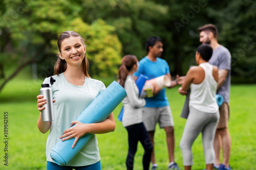 fitness, sport and healthy lifestyle concept - happy smiling young woman with mat and bottle over group of people meeting for yoga class at summer park