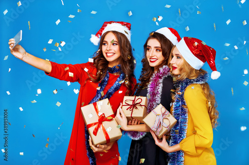 Three smiling women in santa helper hats with many gift boxes make selfie. Young women posing on blue background. Christmas, new year, winter, party concept.
