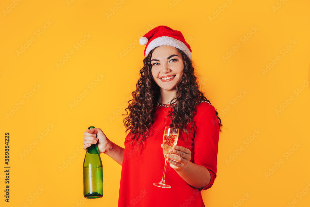 Young beautiful woman on yellow background, on her head she has santa hat. Woman holding bottle and glass in her hands. Christmas mood. Place for text Young beautiful woman on yellow background, on her head she has santa hat. Woman holding bottle and glass in her hands. Christmas mood. Place for text