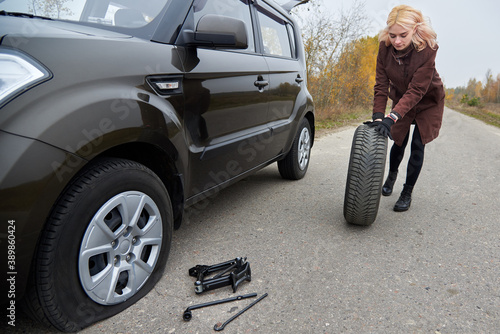 A young blonde woman rolls spare tire near her car with a flat tire.