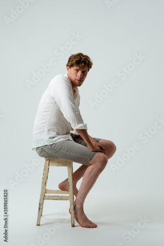 Barefoot man full height sitting on chair. Side view of young guy in white shirt and jeans on white background. High quality photo