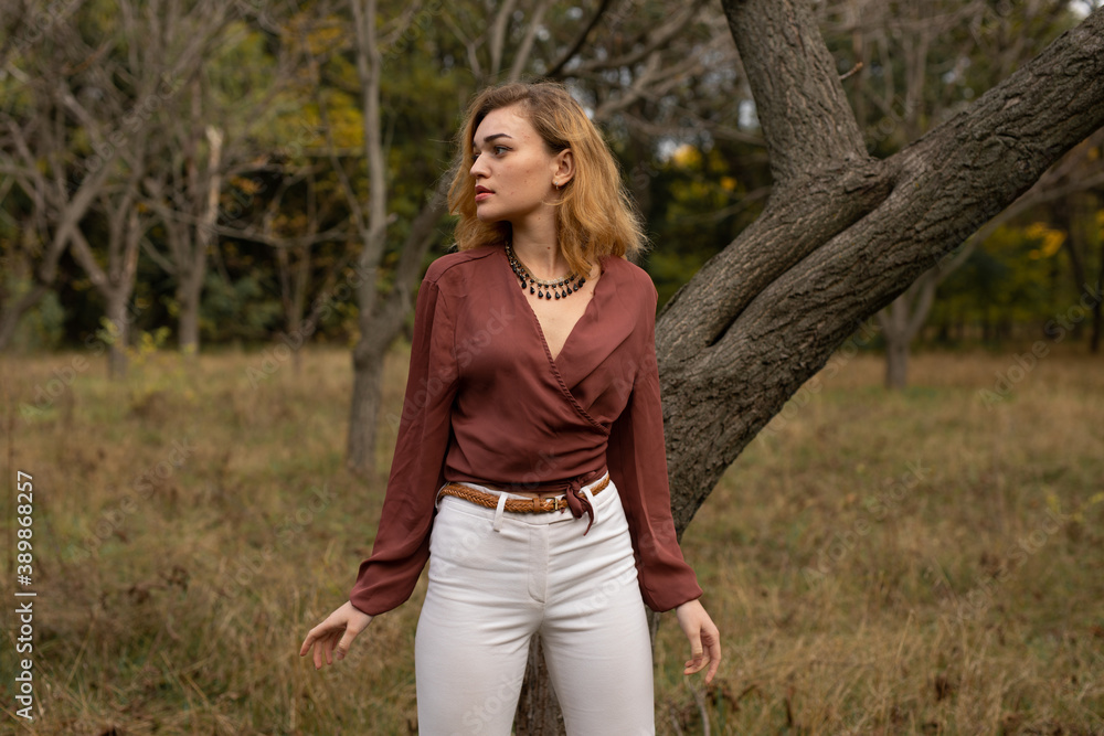 Beautiful blonde woman in in a brown blouse and white trousers posing in the forest Beautiful blonde woman in in a brown blouse and white trousers posing in the forest