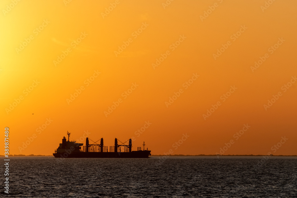 Big cargo ship going through Uruguay river canal at dusk over an orange sky in Conchillas, Colonia, Uruguay. Image with horizontal orientation