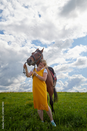 Girl in yellow dress with a horse on a green field and a blue sky with white clouds on the background