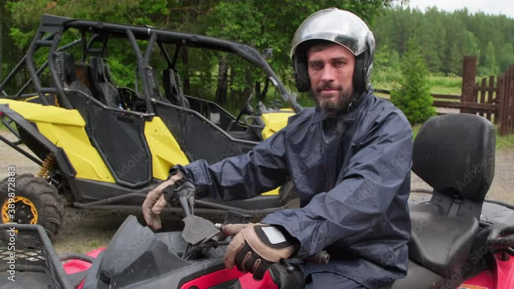 Tracking portrait shot of middle-aged man in helmet and riding gloves sitting on red quad bike and posing for camera outdoors Tracking portrait shot of middle-aged man in helmet and riding gloves sitting on red quad bike and posing for camera outdoors
