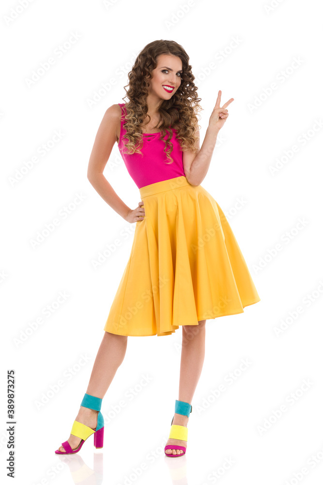 Young Woman In Colorful High Heels, Yellow Skirt And Pink Top Is Showing Peace Hand Sign.