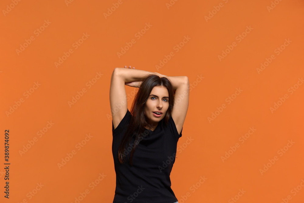 Relaxed carefree caucasian woman in black t-shirt keeps hands behind head, has positive expression, smiles pleasantly, listens something with interest. Isolated over an orange background. Relaxed carefree caucasian woman in black t-shirt keeps hands behind head, has positive expression, smiles pleasantly, listens something with interest. Isolated over an orange background.