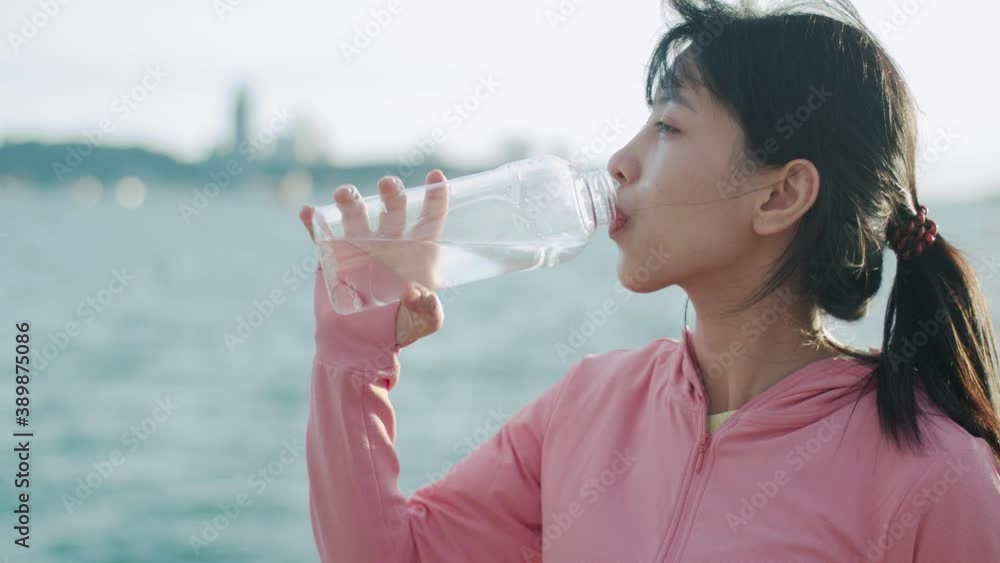Close up of Asian woman runner drinking water while standing seaside. Girl in sports clothes taking a break from exercising admiring the view of the beach sunset. Close up of Asian woman runner drinking water while standing seaside. Girl in sports clothes taking a break from exercising admiring the view of the beach sunset.