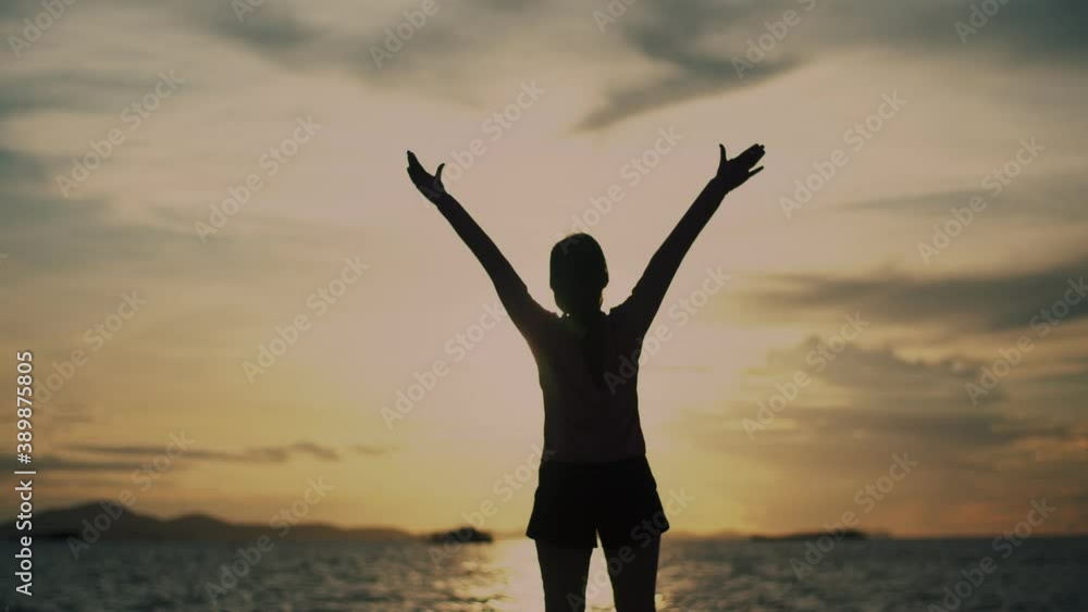Silhouette of a fitness woman in sportswear raises arms into the air looking at the ocean view during a beautiful golden sunset. Silhouette of a fitness woman in sportswear raises arms into the air looking at the ocean view during a beautiful golden sunset.