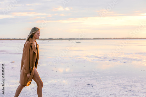 Beautiful young blonde girl with long hair walks along the lake shore and looks at the sunset. Autumn walk. The girl in the coat
