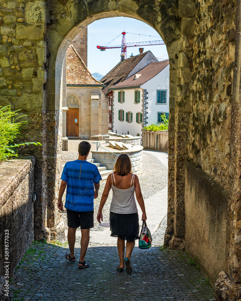 Deux touristes de dos passant sous le portche d'un village ensoleillé avec une place et de maison ainsi qu'une grue avec un ciel bleu l'été en Suisse